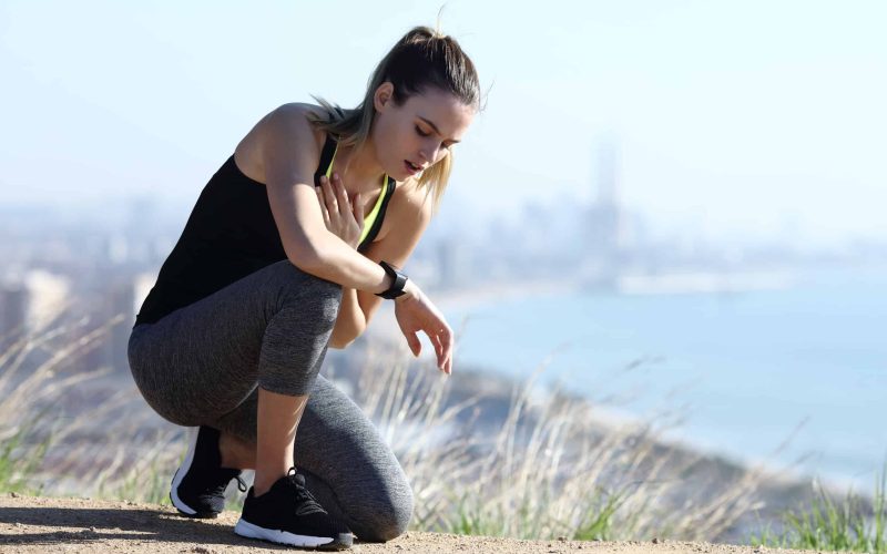 Exhausted suffocated jogger checking pulsations on smartwatch and touching chest outdoors in a city outskirts