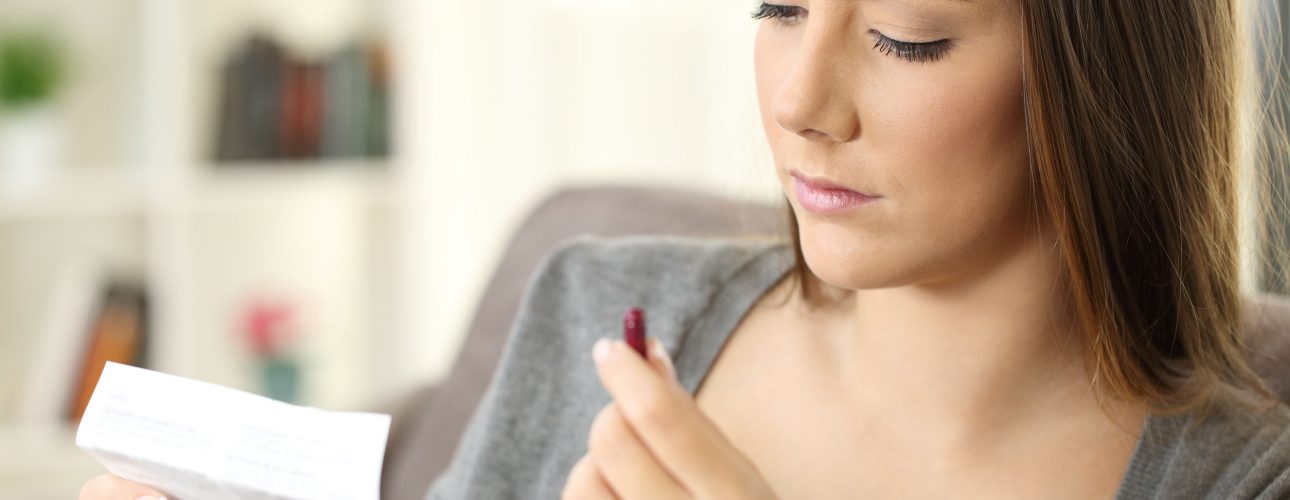 Serious woman holding a medicine capsule reading a leaflet sitting on a sofa in a house interior