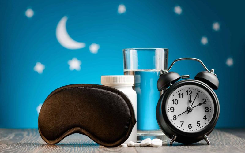 Eye mask, pills and alarm clock on a bedside table over night sky. Getting rid of insomnia, remedies for good restful sleep.