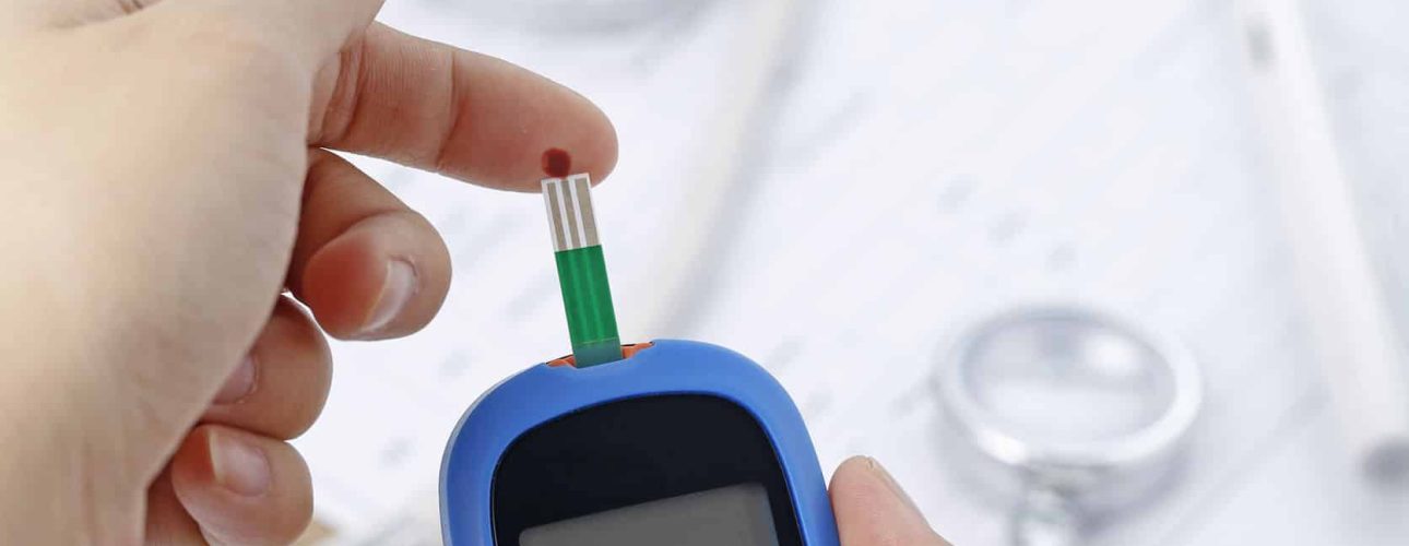 Hand holding a blood glucose meter measuring blood sugar, the background is a stethoscope and chart file