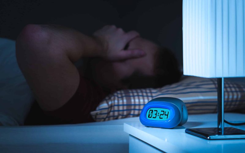 Insomnia and sleepless concept. Man unable to sleep. Exhausted and tired. Covering face with hand. Alarm clock on nightstand and bed in bedroom.