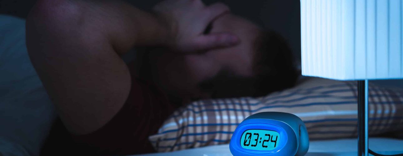 Insomnia and sleepless concept. Man unable to sleep. Exhausted and tired. Covering face with hand. Alarm clock on nightstand and bed in bedroom.